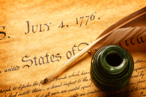 A feather quill and inkwell sitting on top of the American Declaration of Independence. The quill and inkwell sit next to the scribing of one of the most famous dates in world history, July 4, 1776.  A warm golden color scheme dominate the image.