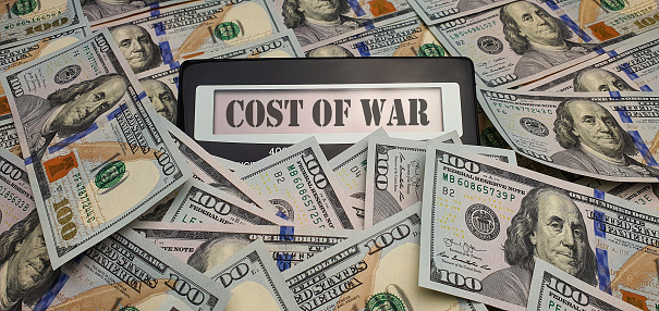 A calculator with the words Cost of War on the screen surrounded by 100 US dollar bills. The concept of global militarization, increasing spending on the army, weapons for defense, fighting terrorism