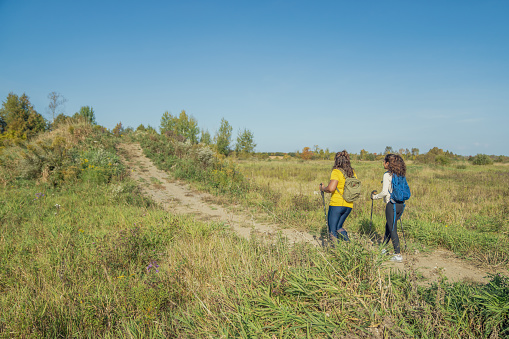 A woman and her daughter hike together on a warm fall day.  They are both dressed comfortably, have backpacks on and hiking poles in hand as they navigate the trails together.
