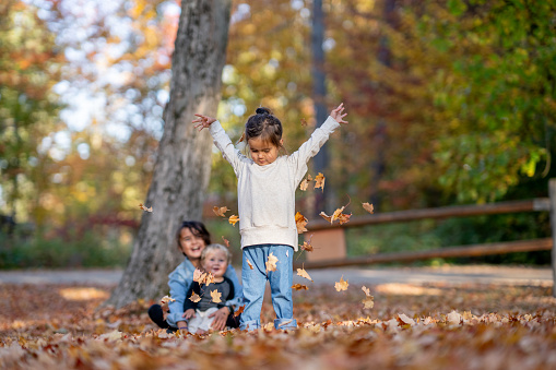 Three siblings play in the fall leaves together during a family camping trip.  They are each dressed casually in fall layers and are giggling as the leaves dance in the air and crunch under them.