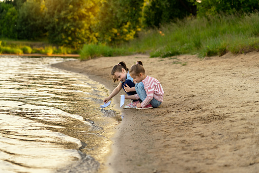 Happy girls play with paper boats on river in green summer park at sunset. Active childrens leisure games outdoors by water