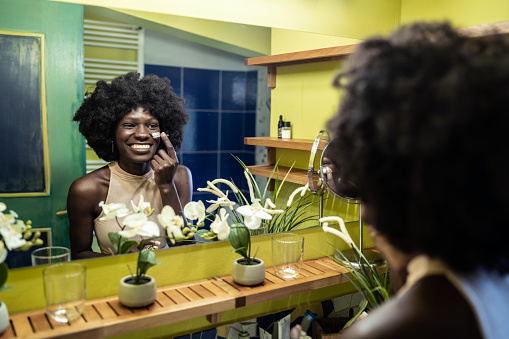 A young  Afro-Caribbean woman is using her finger to spread moisterizer onto her cheek looking into the bathroom mirror