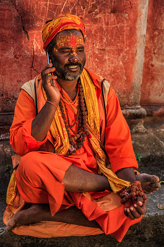 Sadhu - indian holyman using the mobile phone. In Hinduism, sadhu, or shadhu is a common term for a mystic, an ascetic, practitioner of yoga (yogi) and/or wandering monks. The sadhu is solely dedicated to achieving the fourth and final Hindu goal of life, moksha (liberation), through meditation and contemplation of Brahman. Sadhus often wear ochre-colored clothing, symbolizing renunciation.