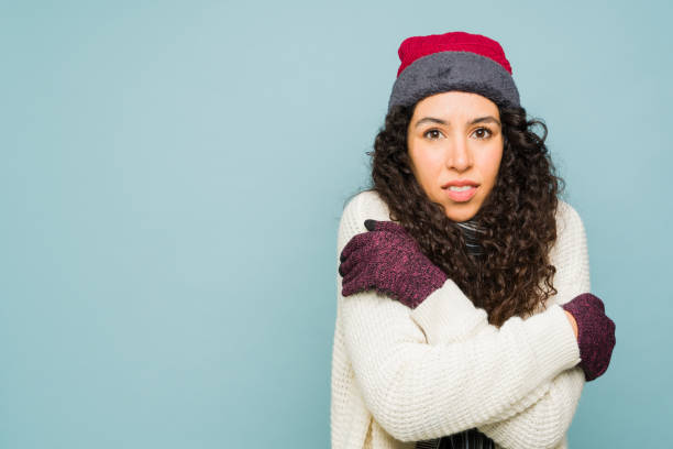 Attractive hispanic woman shivering feeling very cold during winter weather wearing a knit hat, gloves and sweater in a blue background