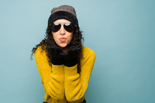 Beautiful young woman with gloves and a knit hat enjoying the cold winter weather blowing a kiss while wearing sunglasses