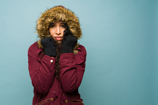 Sad worried young woman putting on a big winter jacket feeling very cold during the winter weather next to copy space ad in a blue background