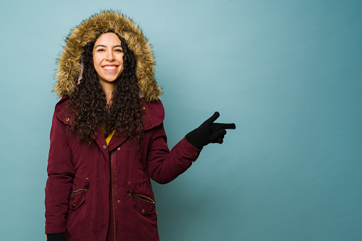 Latin beautiful young woman with a winter jacket enjoying the winter cold weather pointing to blue copy space ad