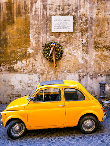 Rome, Italy, March 13 -- A well-preserved yellow model of the famous 1969 Fiat 500 parked along an alley in the Rione Monti, in the heart of the historic center of Rome, under a commemorative marble plaque in memory of the victims of Nazi-fascism in WWII in the concentration camps. In 1980 the historic center of Rome was declared a World Heritage Site by Unesco. Wide angle image in high definition quality.