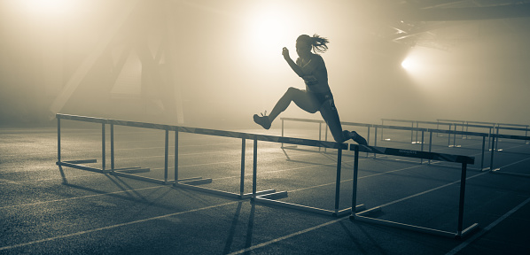 Silhouette of a young female sprinter jumping over hurdles. Sport and competition concept.
