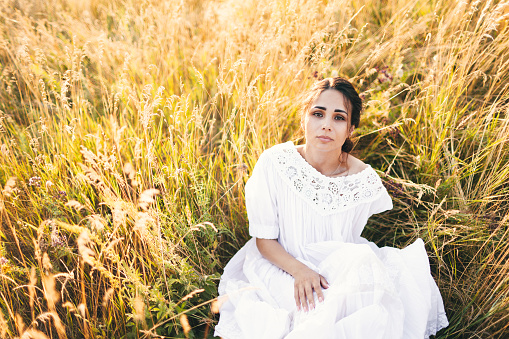 Beautiful girl in white dress sitting in wheat field at sunset