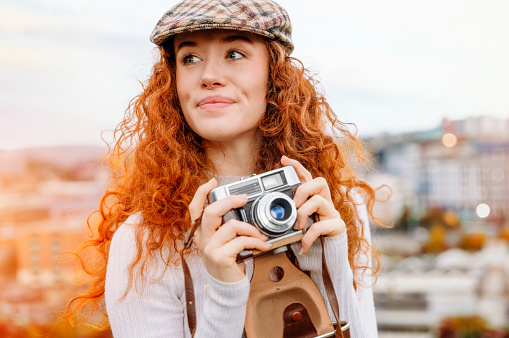 enthusiastic readhead female model in classic English cap laughs happily enjoys spare time taking photo with old fashion camera outside against blurred city background. Lifestyle concept