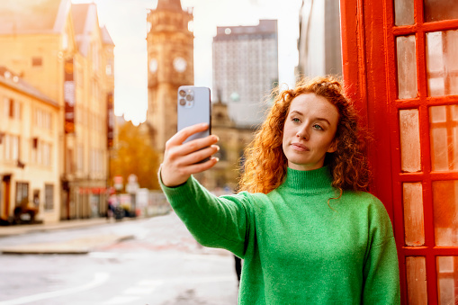 Young woman portrait close to red telephone box in English city taking a selfie with smart phone.  Old city  in the background.