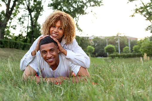 african american young couple in love lies together on the grass in the park in the summer and smiles, curly girl lies on her boyfriend's back and rejoices, copy space