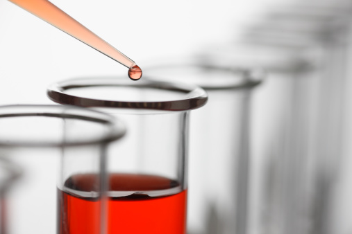 A scientist uses pipette to drop a red solution into a test tube. The test tubes stand in a long row and gently fall out of focus the further they are away from the camera. The neutral color of the test tubes contrasts against the red liquid in the pipette and test tube.