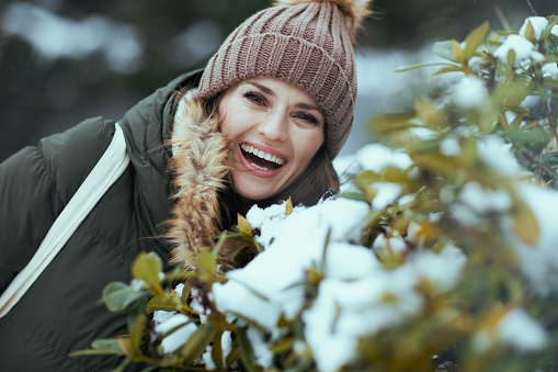 happy modern woman in green coat and brown hat outdoors in the city park in winter with beanie hat near snowy branches.