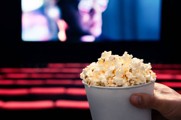 Movies and popcorn. Man holding pop corn box at cinema. Action, thriller or scifi entertainment on screen. Red seats in dark theater. Salty snack in bucket. Spectator pov. stock photo