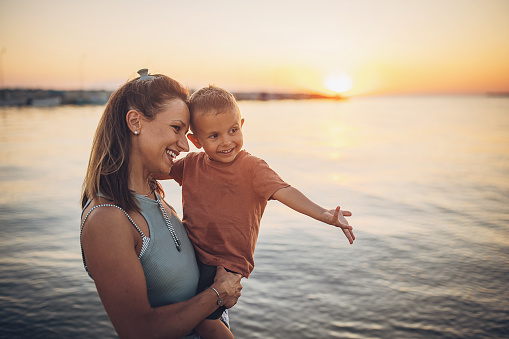 Woman holding her little son while standing on the beach by the sea in sunset, they are on vacation.