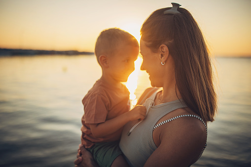 Woman holding her little son while standing on the beach by the sea in sunset, they are on vacation.