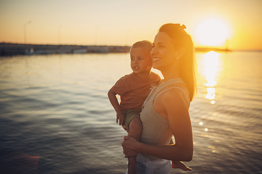 Woman holding her little son while standing on the beach by the sea in sunset, they are on vacation.