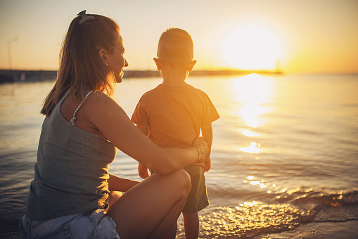 Woman with her little son on the beach by the sea in sunset, they are on vacation.