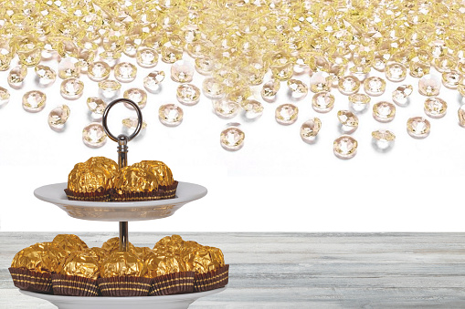 Pastries, desserts and sweets. Chocolate candies wrapped in golden foil on a two-storey etagere on a wooden table. Abstract bright background. Space for design.