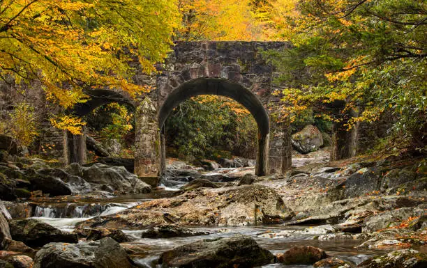 The Triple Arch Bridge, an iconic historic landmark, gracefully spans the Pigeon River in North Carolina along NC-215. This picturesque bridge, constructed in a bygone era, boasts a magnificent trio of stone arches that add architectural grandeur to the rustic landscape. As the golden hues of autumn envelop the river, the Triple Arch Bridge becomes a timeless symbol of nature's beauty and human ingenuity. Visitors are drawn to this charming site to witness the confluence of history, engineering, and the vibrant colors of fall. Explore this enchanting location, where time seems to stand still, and history whispers through the breeze.