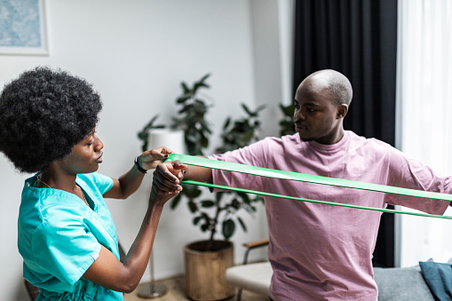 Young black physiotherapist working with patient at home doing exercises with exercise band, helping to recover after surgery