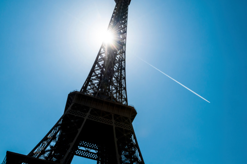 Bottom view of the Eiffel Tower shot against the sun. White airplane track visible in the background.