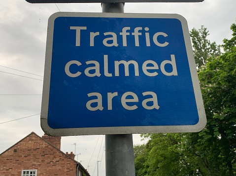 Blue and white rectangular Traffic Calmed area sign, tree and residential house in background UK