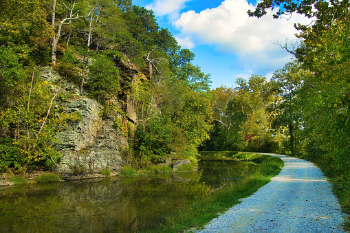 Landscape of an Autumn day on the C&O Canal Towpath Trail in Maryland passing by calm waters and a rocky cliff with colorful trees.