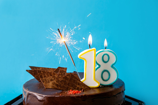 Number 18 birthday candle burning on cake, sparkler sparks fly. The eighteenth birthday or anniversary celebration concept. Lot of copy space on blue background.