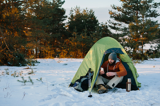 A tired tourist rests after a difficult climb sitting in a tent in a snowy forest. A man uses his phone to send a message or check his GPS.