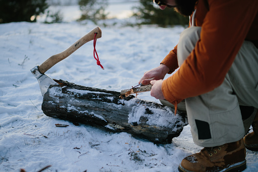 A man prepares wood wool with a knife for starting a fire in a winter forest at sunset. Winter survival concept.