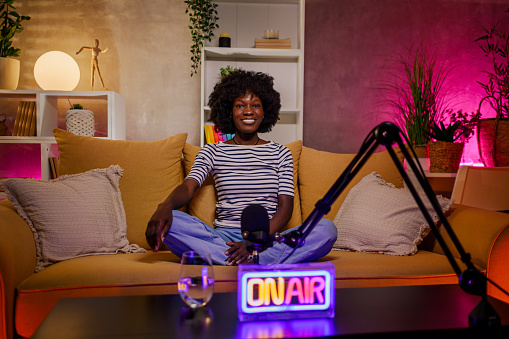 African american woman host streaming her audio podcast and using microphones at her small broadcast studio. black woman radio presenter posing while smiling and looking into the camera. Copy space.
