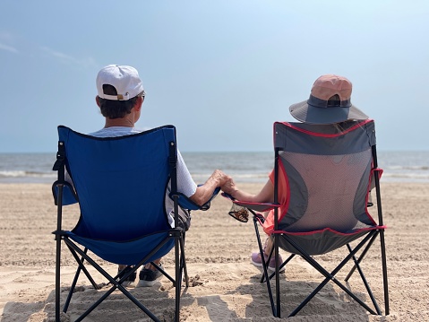 Senior couple, both in hats, sit in chairs holding hands as they look out on Lake Michigan on beach in Two Rivers
