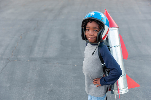 A young boy wears a homemade rocket with flying goggles and helmet while standing on pavement. He is dressed in retro attire ready to fly to the sky.