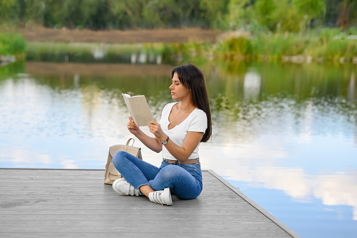 Young woman reading book on pier near lake