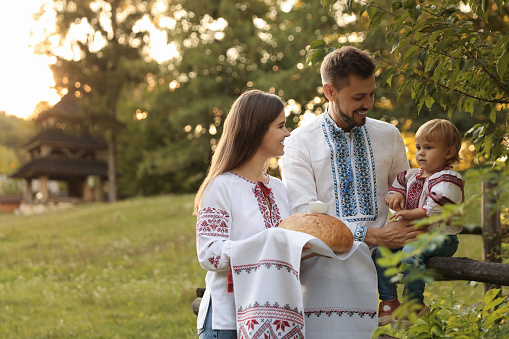 Happy cute family in embroidered Ukrainian shirts with korovai bread near rustic fence outdoors. Space for text