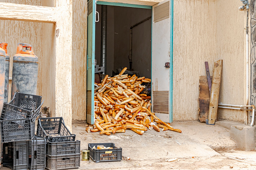 A pile of uneaten bread baguettes thrown away on the ground at the entrance to an old premises with open doors next to gas bottles and crates.