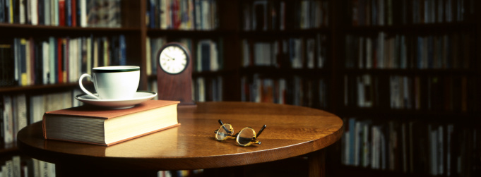 A book, a pair of glasses,a tea cup and a clock on a round table, surrounded by bookcases. The photo is taken with a medium format camera, 80mm lens. Narrow depth of field.