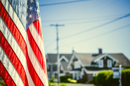 An American flag hanging from a house front porch on the 4th of July is in focus in the foreground, nice homes in the background and a clear blue sky.  Horizontal with copy space.