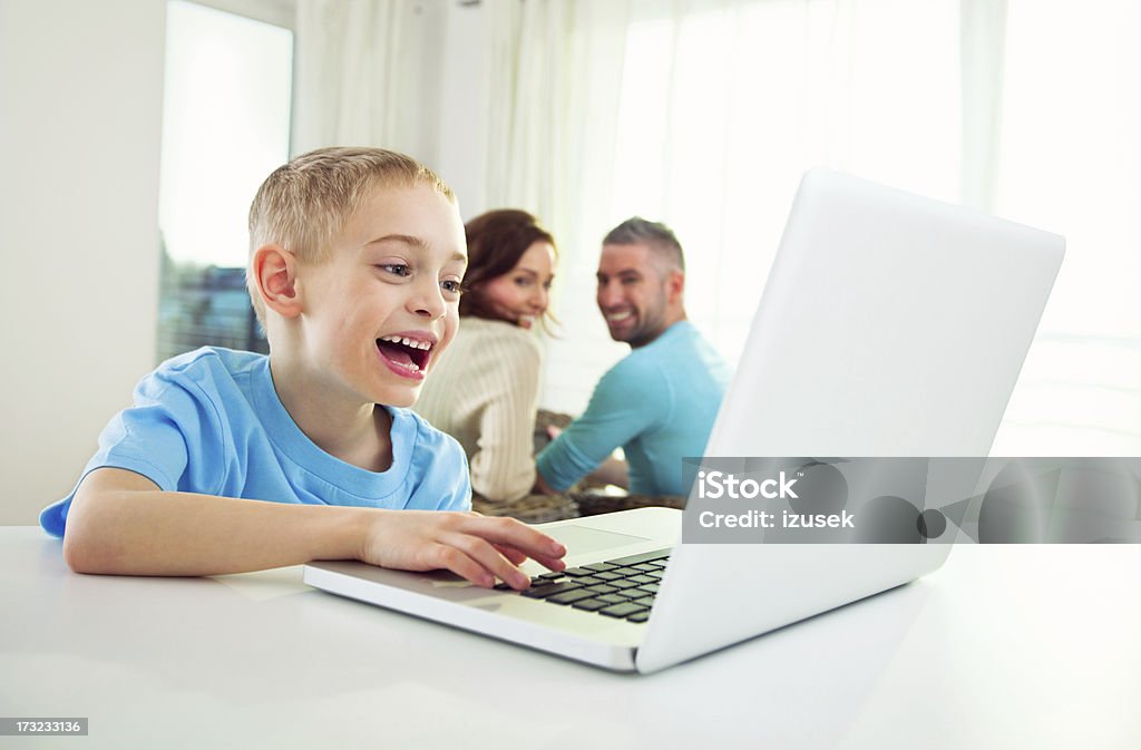 Happy Boy at Home with Laptop Computer A young boy is at home using a large laptop computer and lauging. His parents are in the background smiling and looking at him. 30-34 Years Stock Photo