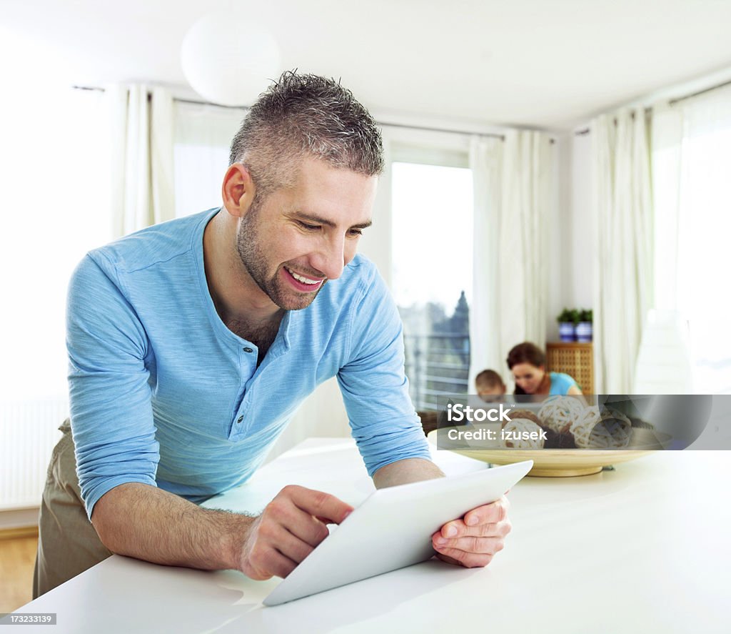 Happy Man with Tablet at Home Close up of a happy man holding a digital tablet and smiling. His wife and son are on the computer in the background. Home Interior Stock Photo