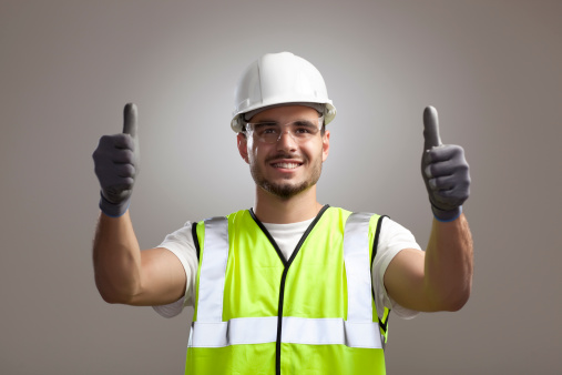 Portrait of happy manual worker man in protective gear (helmet, goggles, gloves) gesturing success sign with both hands up.