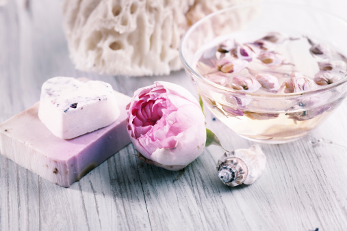 aromatherapy oil in a glass bowl filled with dried roses, a beautiful pink rose and bar of soap in heart shape, all on bright wooden background, image processed in retro style