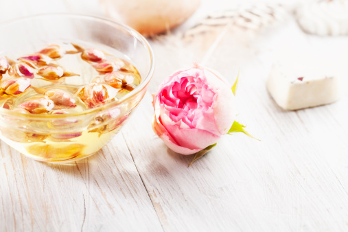 aromatherapy oil in a glass bowl filled with dried roses, a beautiful pink rose, all on bright wooden background