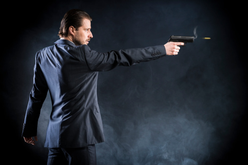 Dangerous man in a suit - assassin, killer, mafia - is aiming with a gun and fires, you can see the smoke from the gun and a speeding bullet. Studio shot, dark background.