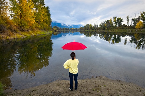 A woman holding a red umbrella stands on the shores by the calm Kootenai River near Bonners Ferry, Idaho.