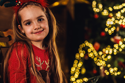 Portrait of beautiful little girl standing by the glistening Christmas tree, smiling at camera with joy.