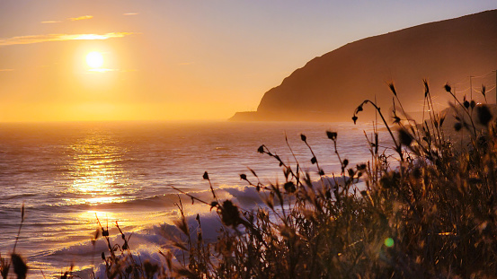 A vibrant orange glow lights up the sky, cutting through the sea mist as the sun approaches the horizon of the Pacific Ocean. Wild grass is silhouetted in the foreground. The view is from the Pacifc Coast Highway near Point Mugu and Malibu, California.